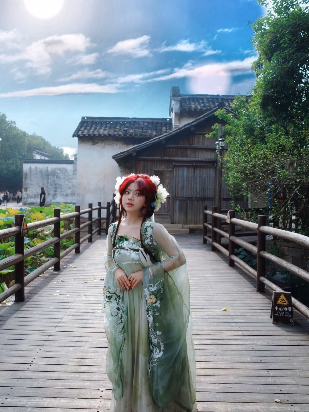 Ánh nắng chiếu nhẹ qua hàng cây, phản chiếu trên mái nhà cổ càng khiến tổng thể trở nên huyền ảo. Bảo Hân đứng giữa lối đi gỗ, dáng vẻ mềm mại và đôi mắt to tròn nhìn xa xăm, khiến người xem liên tưởng đến hình ảnh thiếu nữ thanh thuần trong truyện kiếm hiệp.