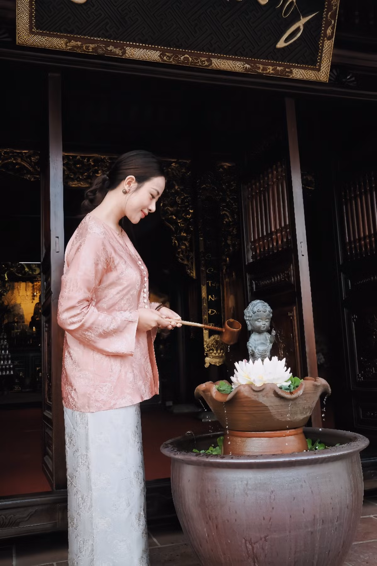 Thiết kế áo dáng truyền thống với họa tiết chìm tinh tế giúp tôn lên vẻ dịu dàng, nền nã, trong khi quần trắng đơn giản tạo cảm giác nhẹ nhàng, thuần khiết. Kiểu tóc búi gọn gàng, lối trang điểm tự nhiên càng làm nổi bật gương mặt thanh tú của bà mẹ hai con.