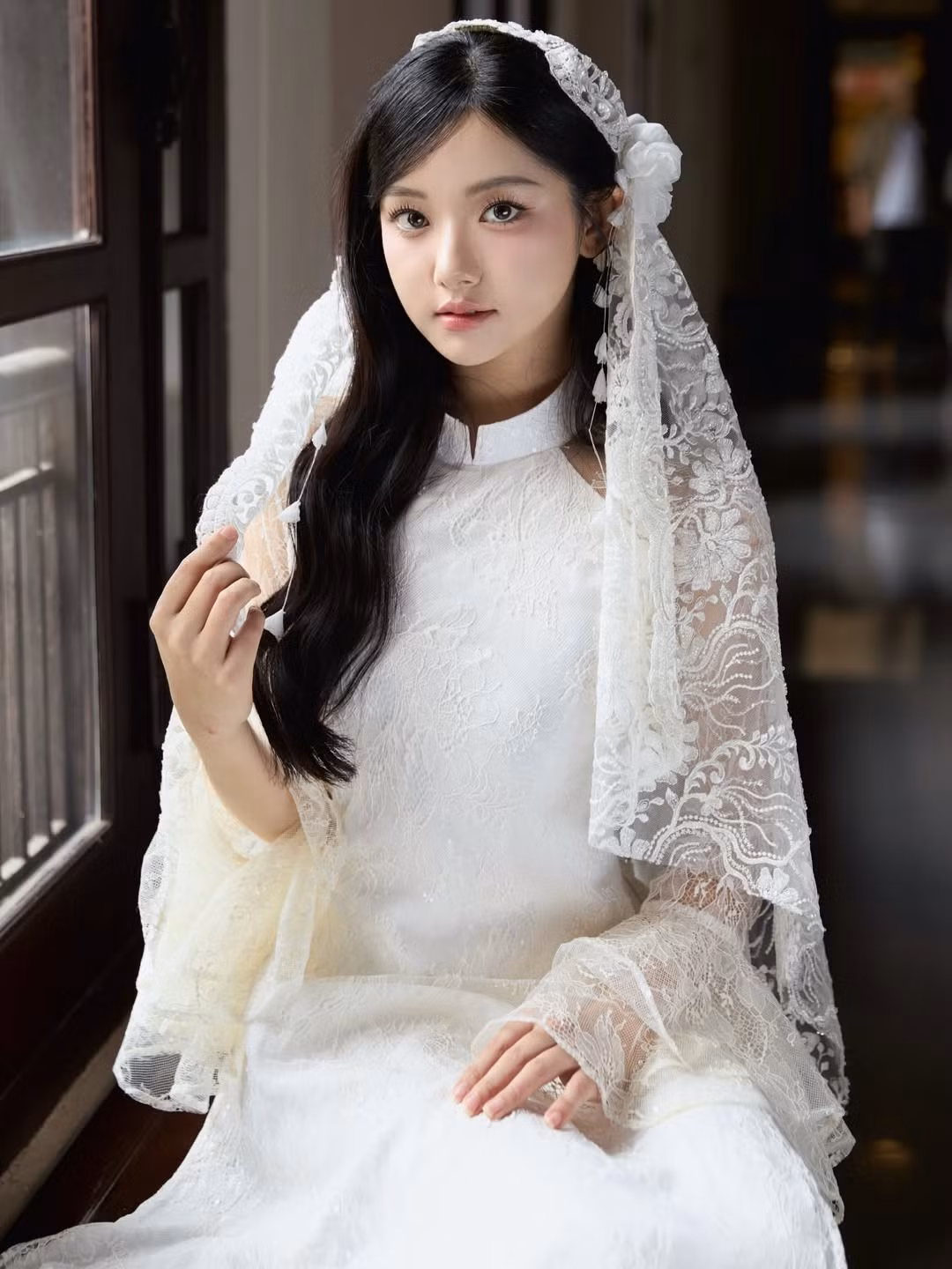 Chiếc áo dài trắng được thiết kế từ chất liệu ren mỏng, tạo hiệu ứng xuyên sáng nhẹ nhàng nhưng vẫn giữ được nét kín đáo cần có. Họa tiết ren hoa chạy dọc thân áo và phần tay áo dài giúp tổng thể thêm phần mềm mại, bay bổng. Phom dáng ôm vừa vặn tôn lên vóc dáng thanh mảnh, trong khi phần cổ cao giữ trọn tinh thần truyền thống của trang phục.