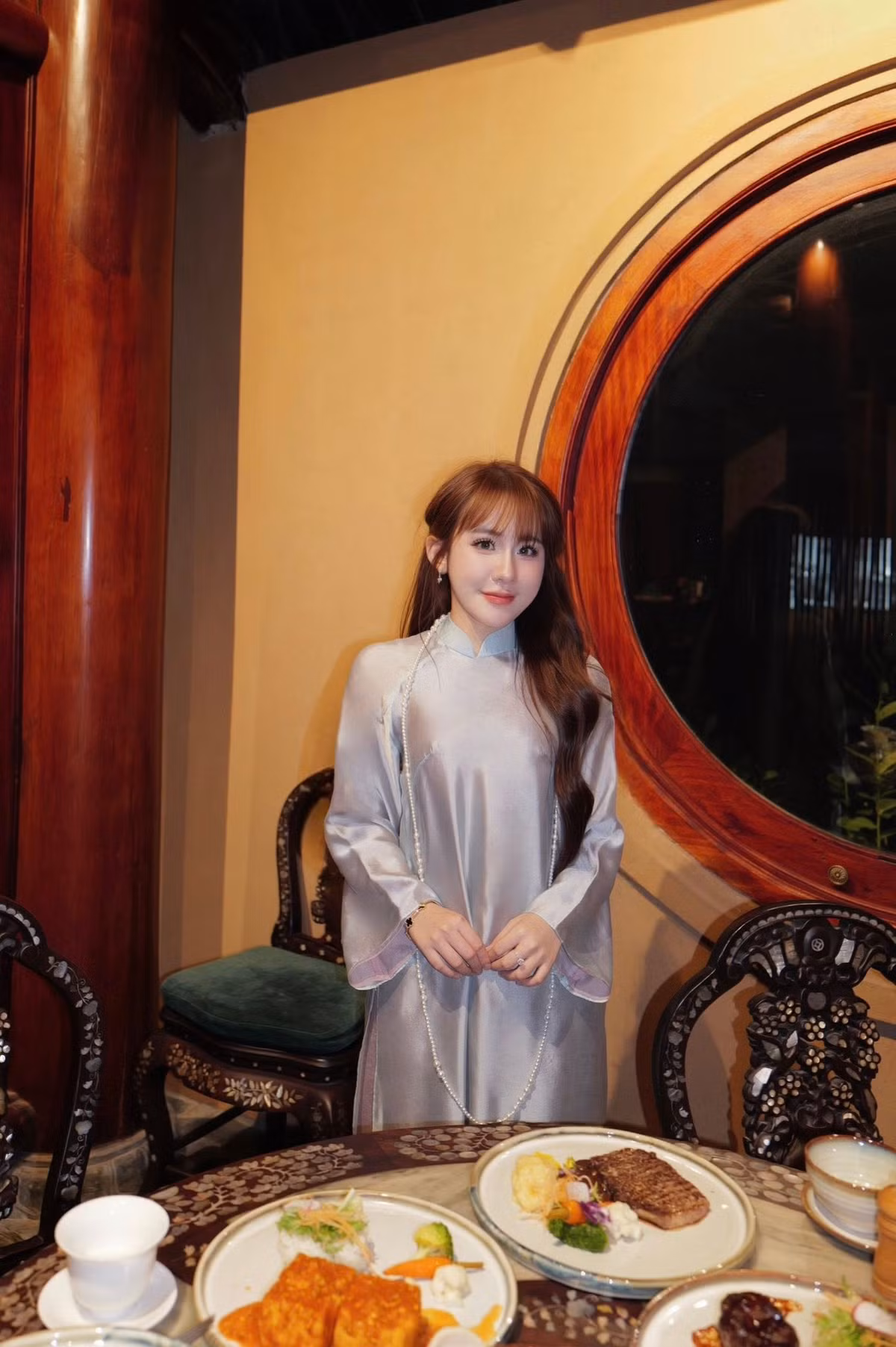 Bối cảnh chụp mang hơi hướng cổ điển với không gian gỗ chạm khắc tinh xảo, bàn trà và các món ăn được bày biện trang nhã. Sự kết hợp giữa trang phục áo dài và không gian truyền thống tạo nên tổng thể hài hòa, giúp bộ ảnh vừa sang trọng vừa gần gũi.