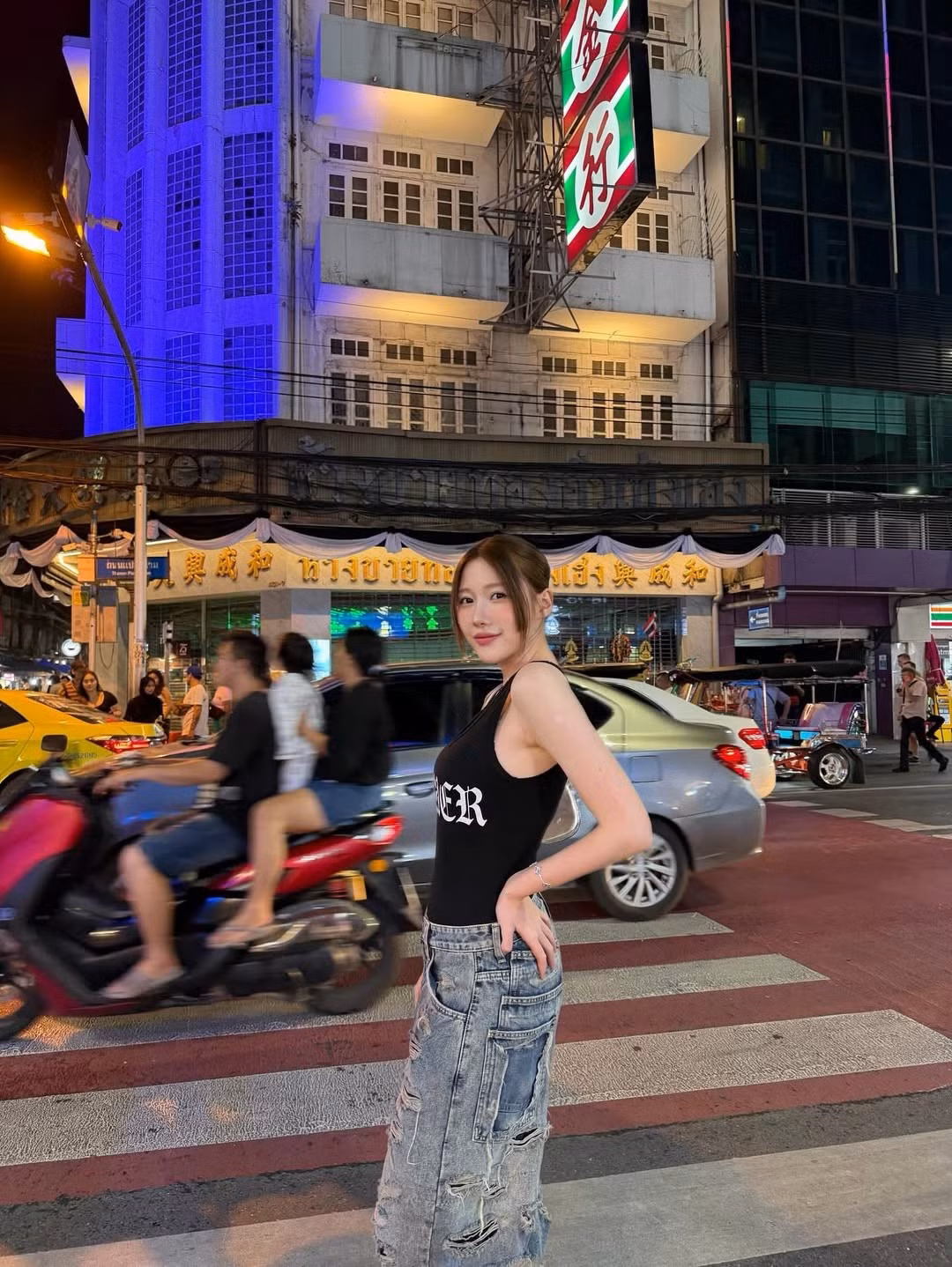 Uyển Ân tự tin tạo dáng "cool ngầu", đút tay vào túi quần, đứng trên vạch sang đường. Bối cảnh phía sau là đường phố Bangkok tấp nập xe cộ, với những chiếc taxi xanh-vàng, xe tuk-tuk và các biển hiệu đặc trưng như "Hotel Royal Bangkok" và "7-Eleven".