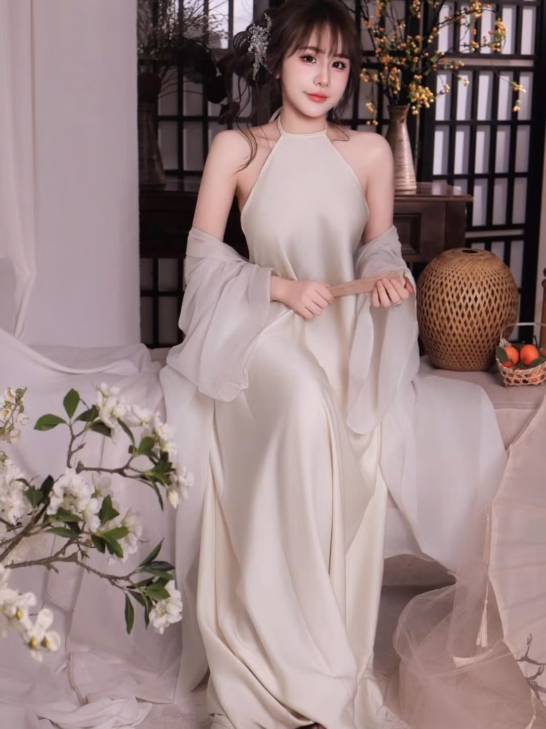 Không gian chụp ảnh cũng góp phần làm nổi bật thần thái của Tuyết Anh. Phông nền mang hơi hướng Á Đông với vách gỗ, bình gốm, cành hoa trắng và ánh sáng dịu nhẹ tạo cảm giác ấm áp, tĩnh lặng.
