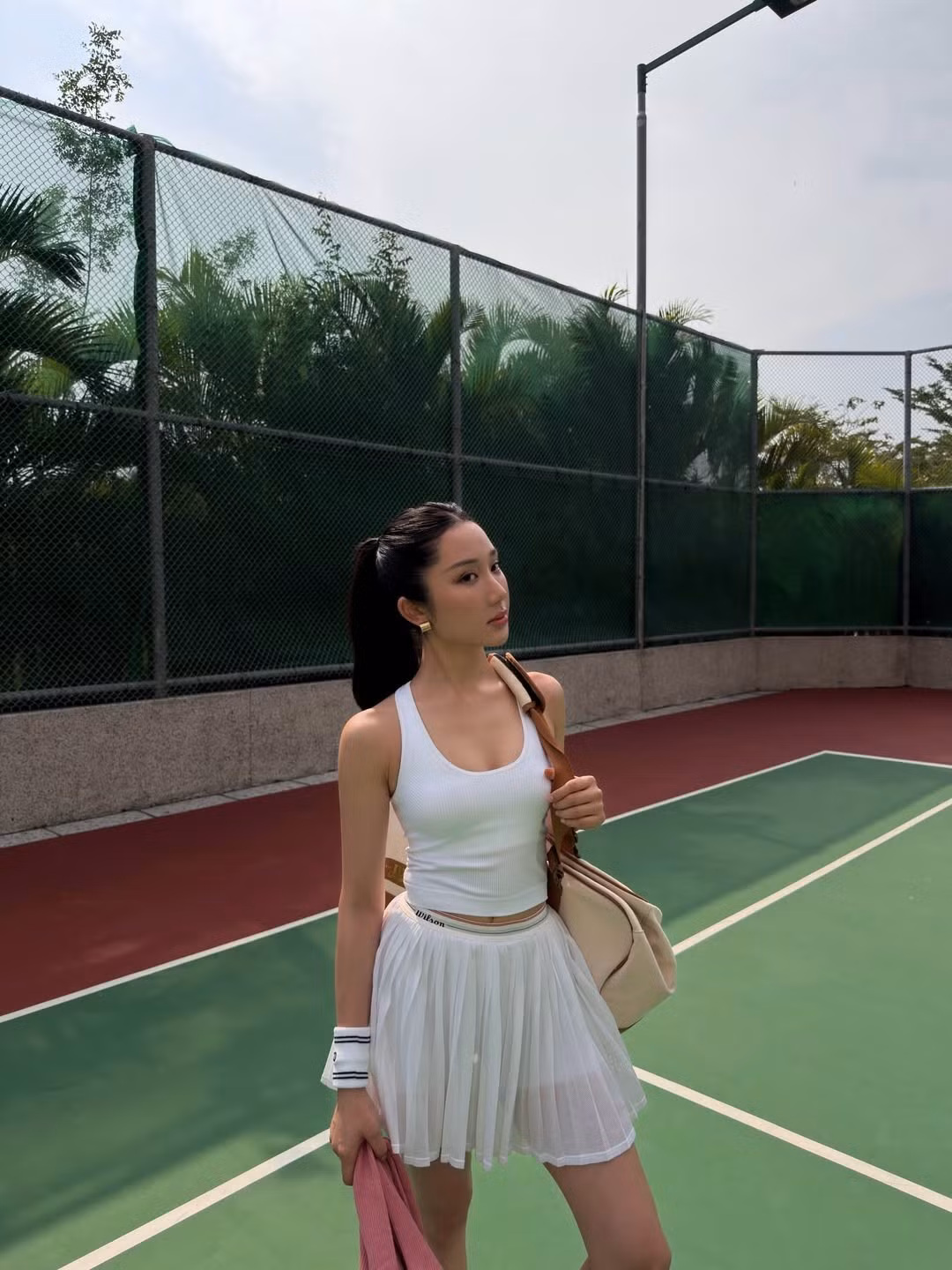 Bối cảnh sân tennis với mặt sân xanh – đỏ đặc trưng, hàng rào lưới và cây xanh phía sau tạo nên không khí thể thao rõ nét. Giữa không gian rộng rãi, ngập nắng, Mina Young thoải mái tạo dáng cùng vợt và bóng tennis, mang lại cảm giác vừa khỏe khoắn vừa thời thượng. Những phụ kiện như túi đựng vợt, khăn, bình nước cũng góp phần làm bộ ảnh thêm phần chân thực, đúng chất một buổi tập luyện thực thụ.