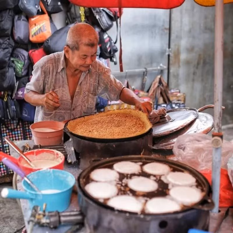 Trên đường Petaling, xe bánh của chú Ah Loh đã đỏ lửa hơn 40 năm với hai món bánh nướng truyền thống Malaysia là apom balik và coconut apom. Apom balik có kích thước lớn, vỏ giòn, ruột mềm dẻo, trong khi coconut apom nhỏ hơn, thơm béo nước cốt dừa. Bột bánh được pha trực tiếp tại xe đẩy và nướng bằng những chiếc khuôn cũ, tạo nên hương vị mộc mạc khó quên. Quán bán từ 13h đến 21h các ngày trong tuần, cuối tuần mở cửa sớm hơn. Ảnh Internet