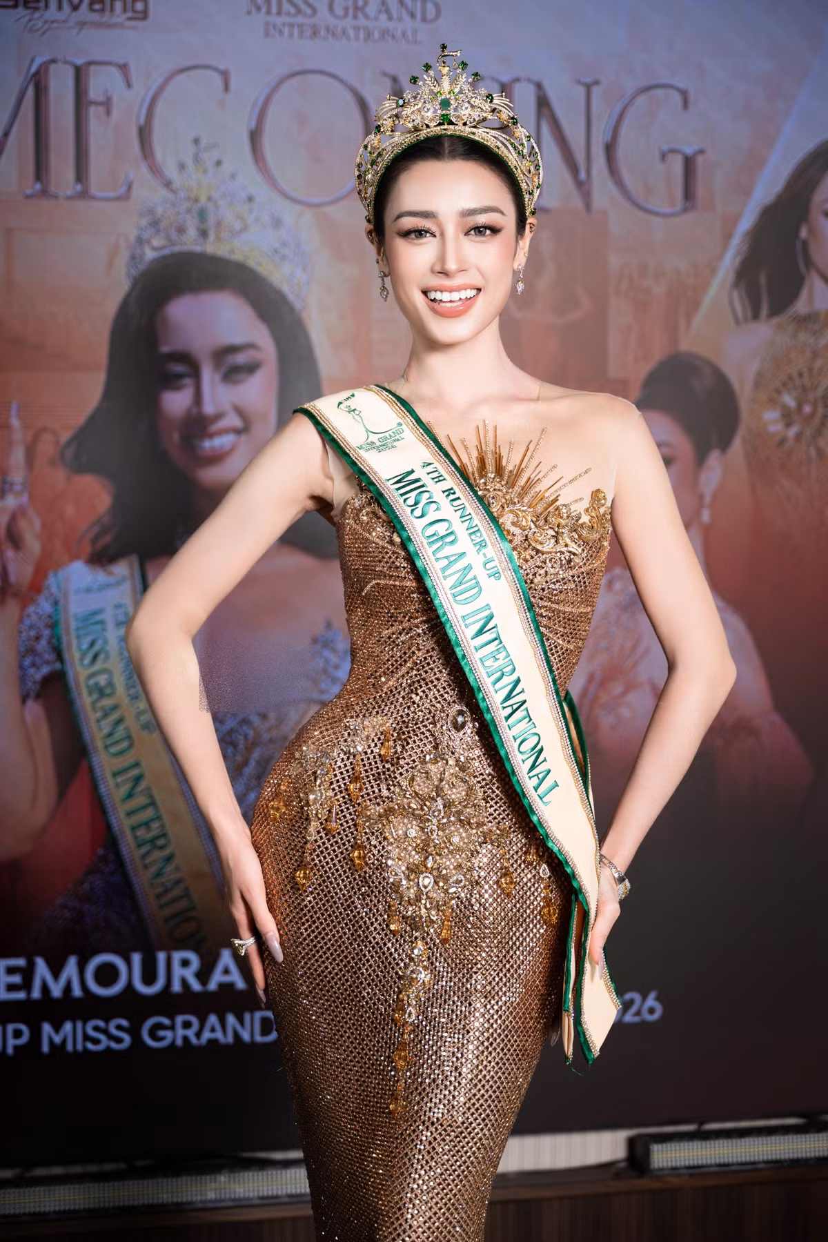Trở về nước, Emoura Phạm xuất hiện trong bộ đầm dạ hội đính kết ánh vàng, đội vương miện và đeo dải băng Á hậu 4 Miss Grand International 2026. Người đẹp khép lại hành trình gần một tháng tranh tài tại Thái Lan với thành tích trong top 5.