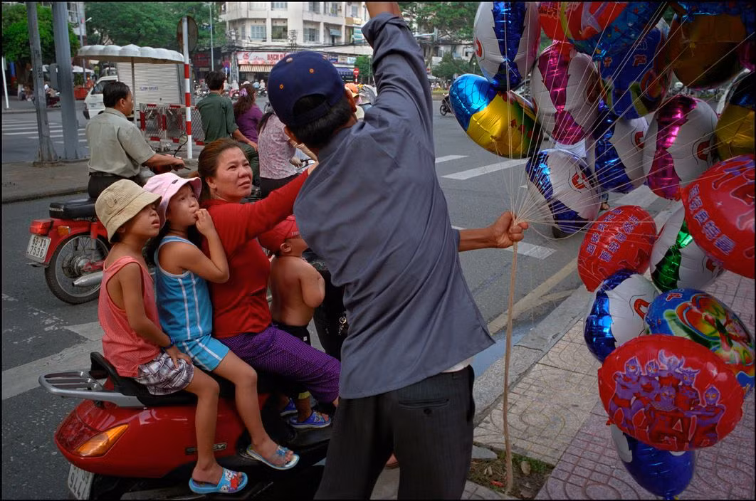 Bà mẹ dừng xe bên đường mua bóng bay cho ba đứa con ngồi trên xe máy. Ảnh: Ian Berry/ Magnum Photos.