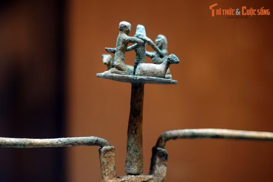 Vẫn là chiếc đèn tìm thấy ở Làng Vạc, trên đỉnh đèn có hình hai người và hai con gia súc, trông giống cừu hoặc dê. Tư thế của hai người giống như đang vận hành một công cụ nào đó. Chi tiết này phản ánh một khía cạnh trong đời sống nông nghiệp của người Việt cổ.