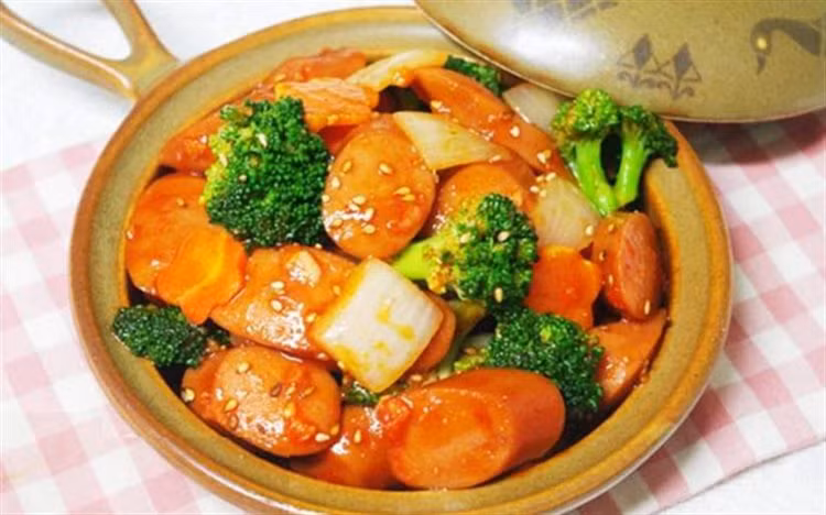 Bắc chảo lên bếp làm nóng chảo rồi cho 2 thìa dầu ăn sau đó cho hành tây, ớt chuông vào đảo đều. Tiếp tục cho xúc xích vào đảo mấy lượt thì cho nước sốt cà, dầu hào và đường vào. Xào đến khi nào nước sốt cạn là được. Ảnh: Cooky.vn.