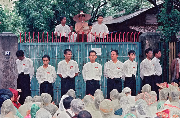Tháng 9/1988, bà chính thức thành lập đảng Liên đoàn Quốc gia vì dân chủ (NLD). Tháng 5/1990, đảng NLD giành chiến thắng trong cuộc bầu cử, nhưng chính quyền quân sự không công nhận kết quả và cáo buộc bà gây chia rẽ quân đội. Do đó, bà chịu sự quản thúc tại gia trong vòng 2 năm. Ảnh: Những người ủng hộ tập trung bên ngoài nhà của bà để nghe diễn thuyết năm 1996.