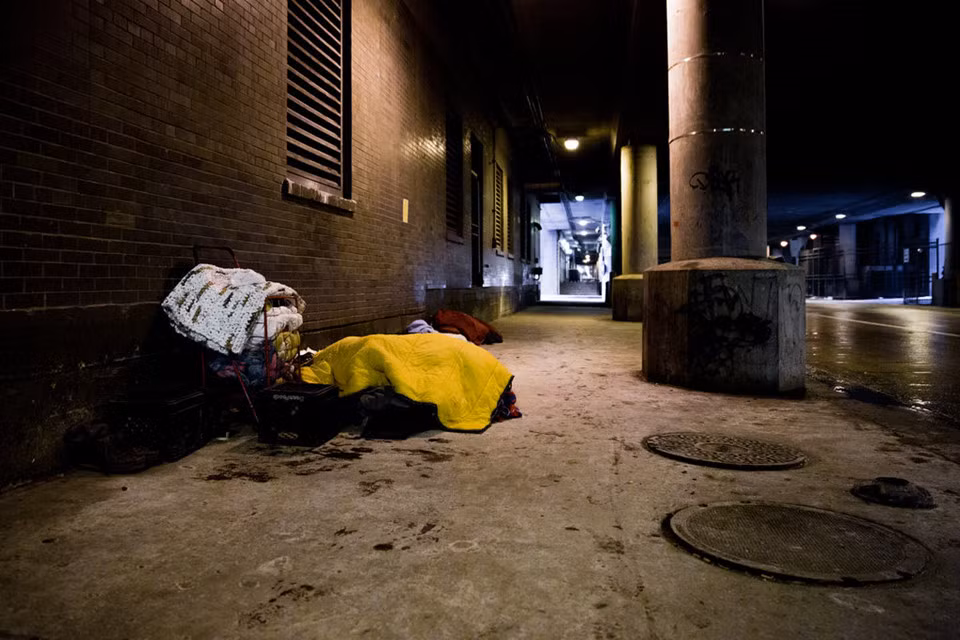 Một khu người vô gia cư thường tới lui ở đường Lower Wacker Drive, Chicago, nơi một người đang ngủ dưới nhiều lớp chăn ở cửa vào một tiệm thuốc. Trong khi người dân thành phố đang gấp rút mua hàng để chuẩn bị cho tình huống xấu nhất, người vô gia cư 60 tuổi này chỉ có thể đeo trên cổ tấm biển xin được giúp đỡ. Ông đã ngủ ngoài đường đêm trước đó, và cơ thể vẫn chưa thể ấm lên. Ông nói chưa biết sẽ ngủ ở đâu đêm nay. Ảnh: New York Times.