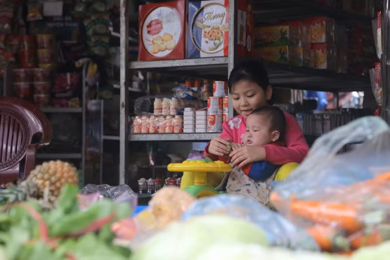 Tại một cửa hàng bách hóa trong thôn, bé gái thay mẹ vừa trông hàng vừa dỗ em để mẹ chuẩn bị bữa cơm trưa.