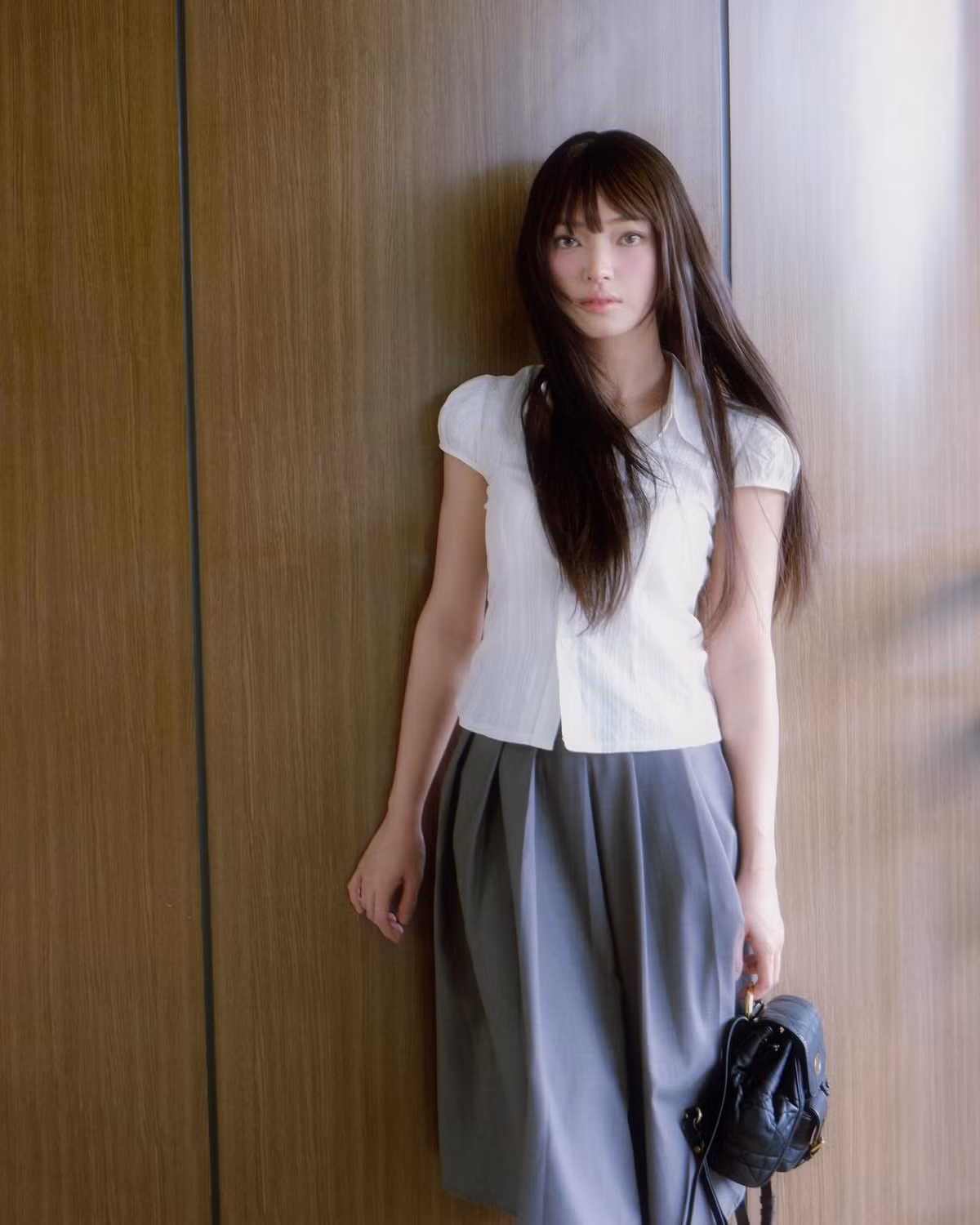 Châu Bùi xuất hiện trong trang phục áo blouse trắng tay bồng dịu dàng, kết hợp với chân váy xếp ly màu ghi dài ngang gối, tạo nên vẻ điệu đà và thanh lịch.