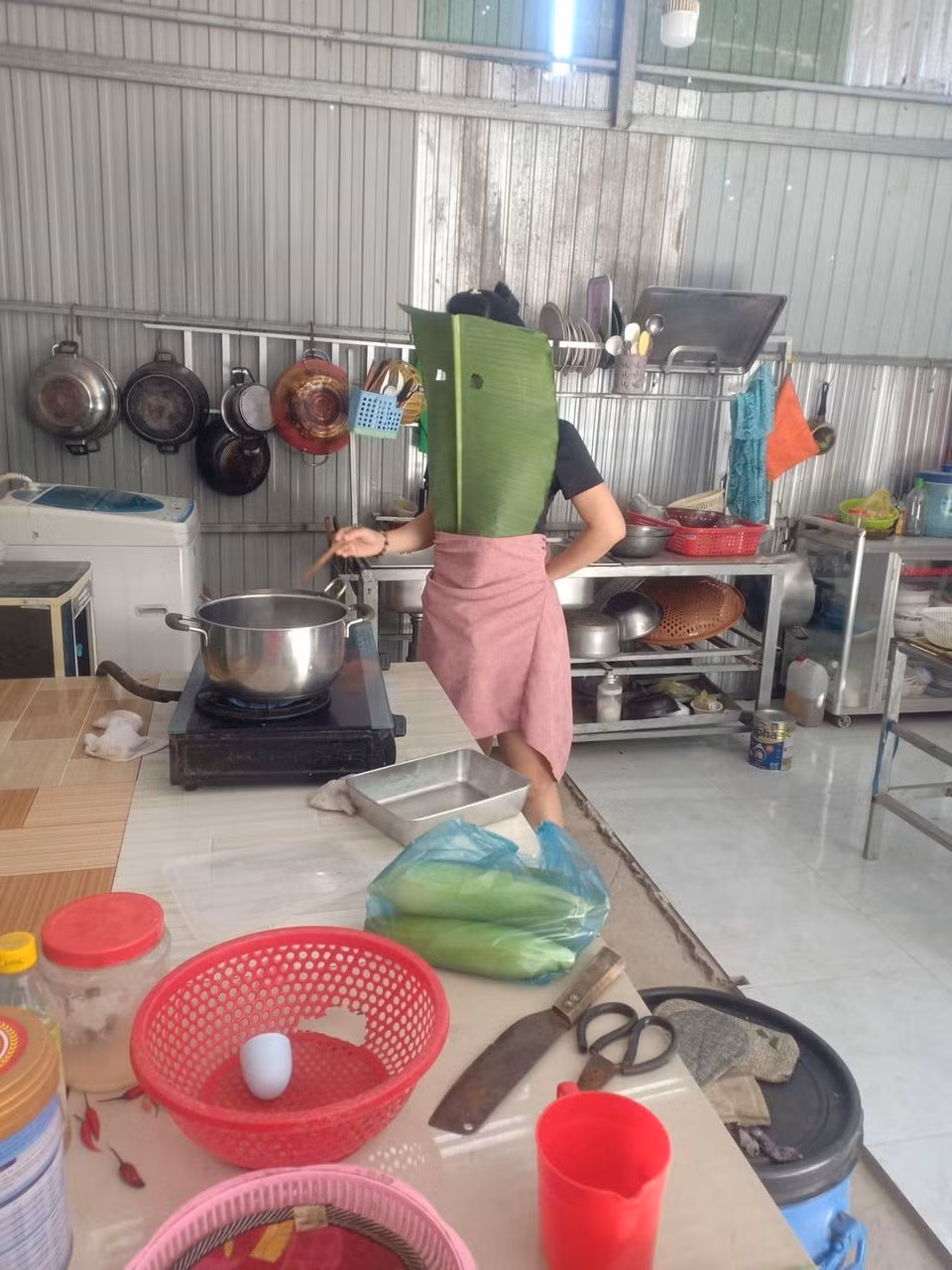 Để chuẩn bị cho màn rán cá không bị bắn của mình, người phụ nữ đã ra vườn, chọn chiếc lá chuối phù hợp sau đó đeo lên người và điều thú vị là không quên khoét 2 cái lỗ để nhìn chảo cá rán.