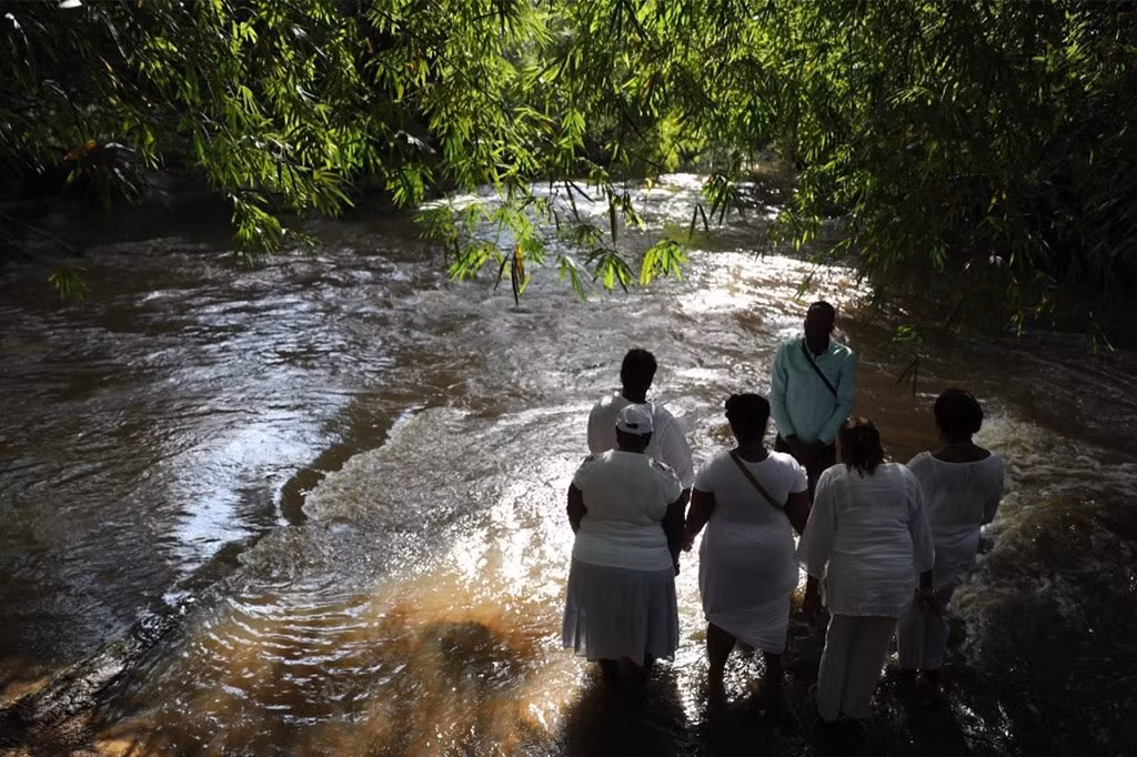 Gần cuối hành trình ở Ghana, những người bị bắt được tắm lần cuối trên một dòng sông như nghi thức trước khi bán. Ngày nay, con sông Assin Manso trở thành nơi tưởng niệm linh thiêng. Trong ảnh, bà Regis Thomson, 75 tuổi, người New York, quây vòng tròn với 5 người phụ nữ khác từ nhà thờ của mình và cầu nguyện trên sông Assin Manso.