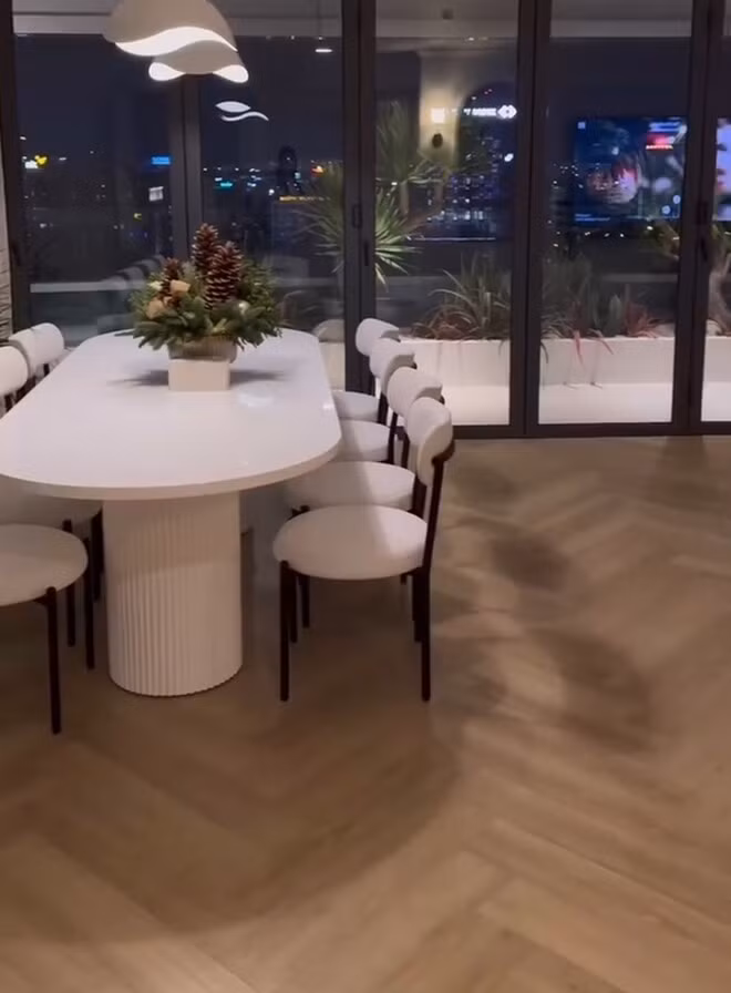 Phòng ăn có view hướng ra thành phố rực rỡ ánh đèn nhờ cửa kính lớn. Trên bàn ăn đặt chậu thông Noel, phía trên là đèn trần nghệ thuật. Ảnh chụp màn hình