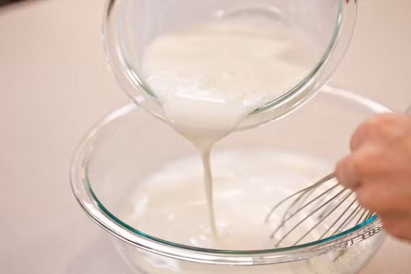 B2. Sau đó bạn cho toàn bộ sữa tươi và lá glatin đã mềm vào trong nồi (khoảng 500 ml) cùng với sữa đặc, sữa milo để lên bếp đun nóng khoảng 45-50độ C và không để hỗn hợp sôi ngoáy thật đều. Bạn đổ sữa chua vào nồi sữa tươi và sữa đặc ở trên sau đó khuấy thật đều để hòa tan hoàn toàn hỗn hợp quyện vào với nhau. Ảnh. Agiadinh.