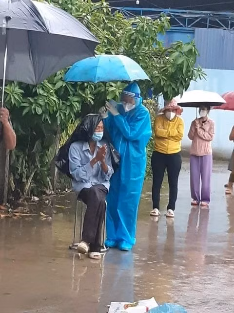Hình ảnh tình nguyện viên che ô tránh mưa cho một người dân đang xếp hàng chờ.