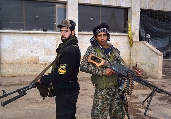Hai chiến binh thuộc phong trào Hezbollah al-Nujaba cầm vũ khí đứng canh gác trước một tòa nhà.