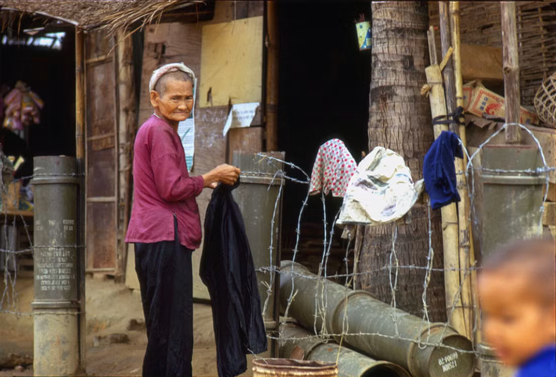 Cụ bà bên ngoài ngôi nhà có hàng rào dựng bằng vỏ bình xăng của quân đội Mỹ. Ảnh: Mark Melnick.