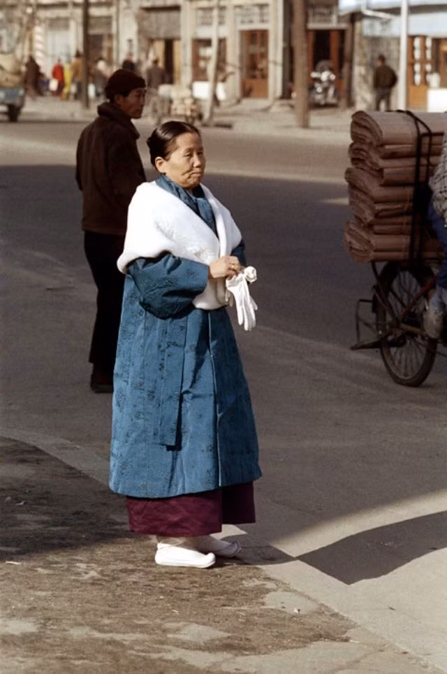 Một phụ nữ lớn tuổi mặc trang phục truyền thống đứng trên vỉa hè tại một con phố đông đúc ở Seoul.