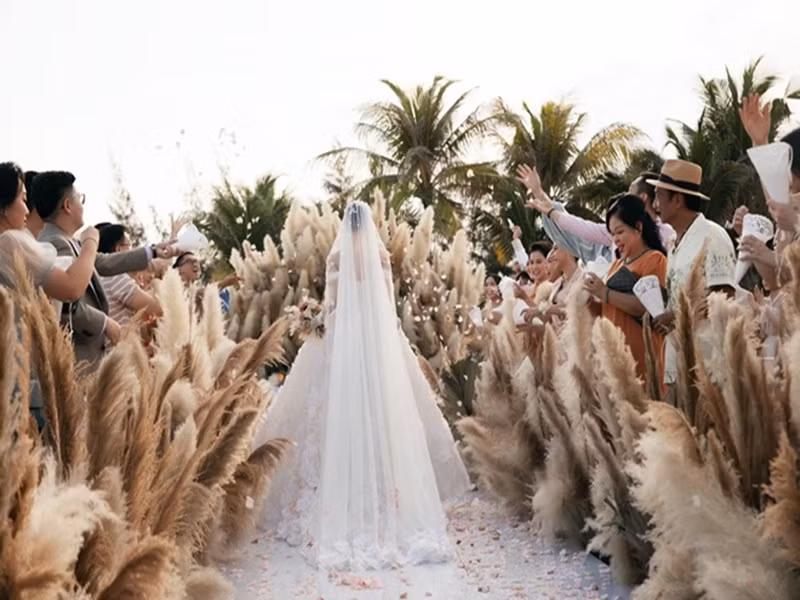Không gian đám cưới của Minh Hằng tràn ngập cỏ lau. "Lý do chọn cỏ lau bắt nguồn từ một trò chơi thuở bé. Tôi muốn mình là bông hoa duy nhất nên đã không chọn bất kỳ một bông hoa nào xuất hiện trên lễ đường. Hằng là bông hoa cho người đàn ông của đời mình. Thật ra cỏ lau là một loài hoa có sức sống vô cùng kiên cường dù trải qua bao bão giông. Dù rất mềm mại, đung đưa nhưng không dễ khuất phục. Tôi muốn tình yêu của mình cũng sẽ như thế. Bên cạnh đó, với việc tổ chức ngoài biển thì Hằng muốn đồ trang trí phải có sự đung đưa, hòa nhịp với gió trò chứ không phải những bông hoa rực rỡ nhưng đứng yên một chỗ. Điều này cũng tượng trưng cho cá tính của cô dâu, rất mạnh mẽ nhưng không kém phần uyển chuyển", Minh Hằng chia sẻ. Ảnh: Người đưa tin