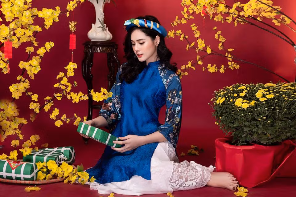 Người đẹp mặc áo dài xanh với váy trắng xinh xắn bên hoa mai vàng và bánh chưng xanh.