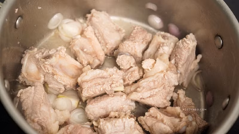 Bước 3: Lấy một cái nồi khác, đổ một lượng dầu thích hợp, cho hành băm nhỏ và gừng thái sợi vào phi thơm, đổ sườn đã chần vào, quấy đều một lúc trên lửa nhỏ. Khi thịt trên bề mặt sườn vàng đều thì đổ một lượng rượu nấu ăn thích hợp vào trộn đều để khử mùi tanh hiệu quả. (Ảnh: cookbeo)