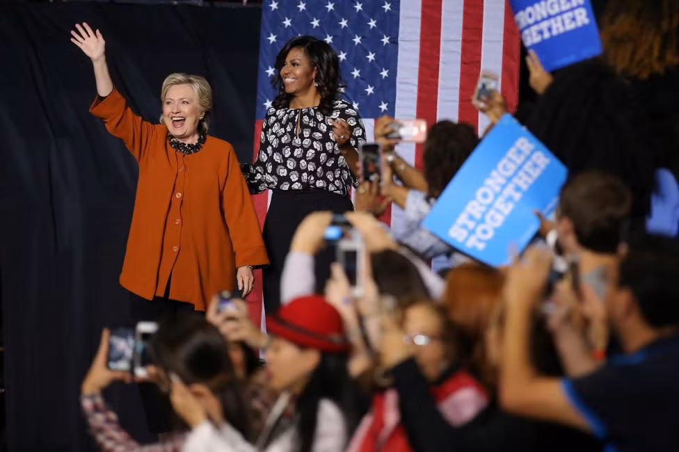 Cựu Ngoại trưởng Mỹ Clinton vẫy tay chào người ủng hộ trong khi bà Michelle Obama đứng cạnh.