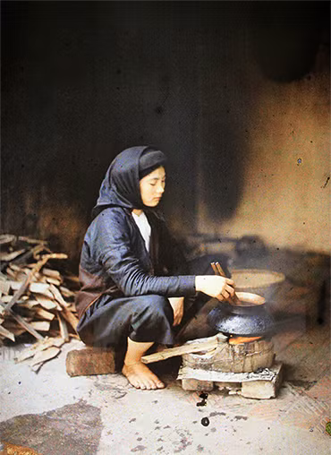 Một phụ nữ đang ngồi nấu cơm với bếp củi. Trông trang phục có thể đoán đây là một phụ nữ thuộc tầng lớp trung lưu.