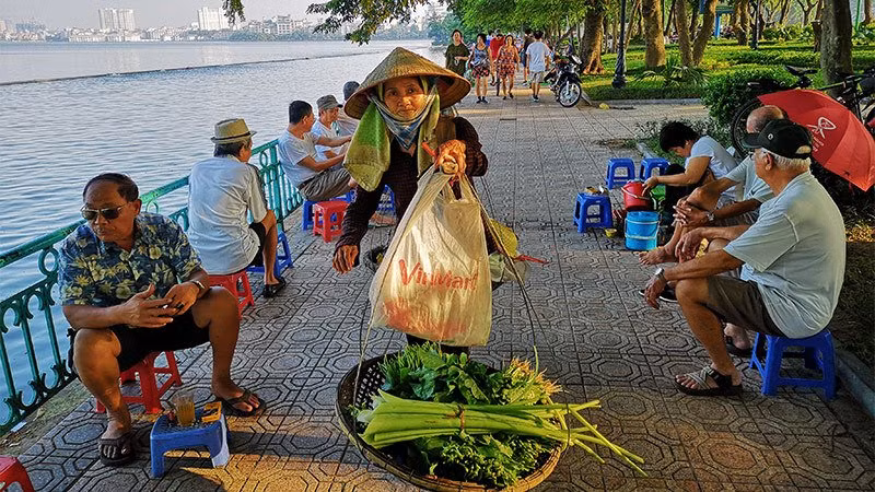 Một phụ nữ gánh hàng rau cho kịp buổi chợ sớm đi qua những người đàn ông đang trầm tư bên chén chè sớm ven Hồ Tây.