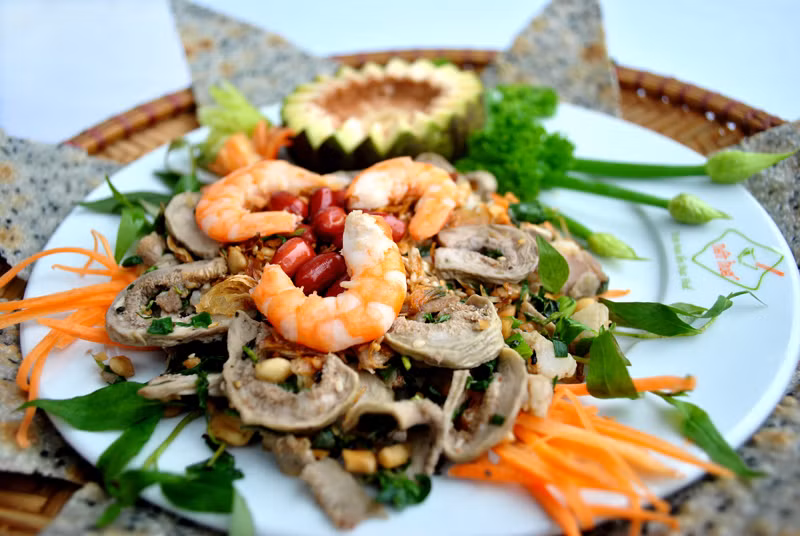 Dùng bao tay trộn tất cả nguyên liệu gồm cả mè và đậu phộng (rau húng quăn cũng được trộn vào, chỉ chừa vài nhánh để trang trí trên mặt đĩa gỏi cho đẹp) cùng chung với phần nước mắm đã chuẩn bị. Sau cùng dọn ra đĩa, rải ớt và rau húng quăn lên. Ảnh: nethue.com.vn.