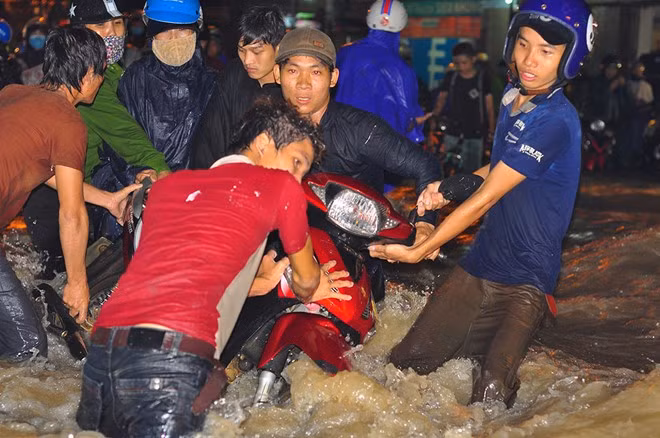 Nước từ các con hẻm cao trong khu dân cư dồn về đường Bùi Văn Hòa (đoạn thuộc khu phố 7, phường Long Bình) tạo thành dòng chảy xiết. Nhiều người cố gắng đẩy xe qua chỗ ngập nhưng bị nước xô ngã, cuốn trôi. Trong ảnh: Một nhóm thanh niên vây quanh chiếc xe máy, giúp chủ phương tiện khiêng qua chỗ ngập.