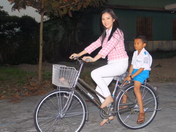 Xuất hiện trong bộ đồ giản dị - áo sơ mi kẻ nhẹ nhàng kết hợp với quần jeans, tóc đen buông xõa, người đẹp đã cởi bỏ giày, đi chân trần để dễ đạp xe đạp khi chở cậu bé học sinh nghèo trên chiếc xe đạp mới là quà tặng mà cô mang đến cho các em nhỏ.