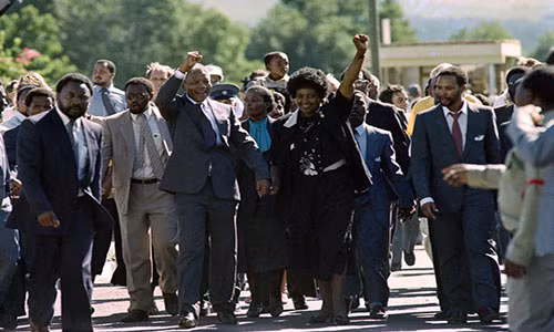 Vợ chồng ông Mandela nắm tay nhau rời khỏi nhà tù Victor Verster và vẫy chào đám đông vào năm 1990 ở Paaarl, Nam Phi. Ảnh: Getty.