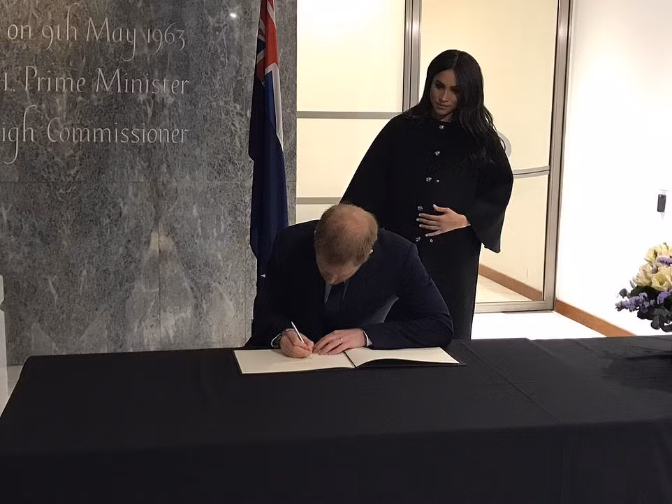 Hoàng tử Harry viết vào sổ tang tại toà nhà New Zealand House ở London trong khi Công nương Meghan Markle đứng phía sau.