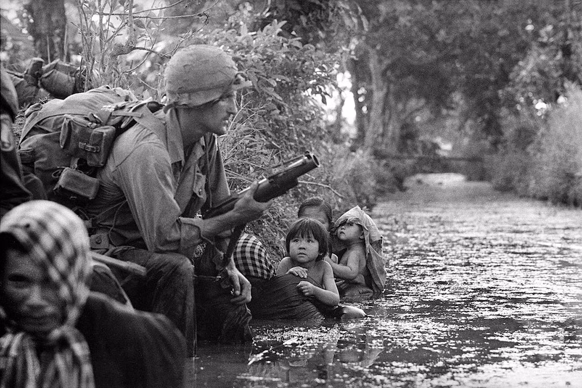 Những đứa trẻ nhìn chằm chằm vào một lính dù Mỹ đang cầm súng phóng lựu M79 trong lúc cùng mẹ ẩn nấp dưới bờ kênh trong chiến sự tại khu vực Bào Trai (nay là Hậu Nghĩa, Long An), phía Tây Sài Gòn, 1/1/1966. Ảnh: Horst Faas/ AP