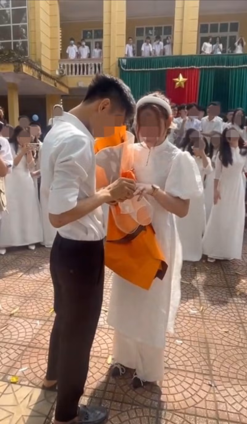 Theo clip ghi lại, ngay sau lễ bế giảng, một nam sinh với đồng phục quần tây - áo sơ mi bảnh bao bước vào khu sân khấu chính với bó hoa lớn trên tay. Đứng trước bạn gái diện áo dài, anh chàng rút trong túi quần một hộp đựng nhẫn và đeo lên tay nữ sinh trong sự reo hò của bạn bè. Ảnh: cắt ra từ clip