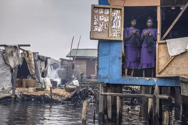 Hình ảnh hai người phụ nữ tại một cửa tiệm làm tóc tại khu ổ chuột ở Makoto, Nigeria. Trước đây, dòng sông này là nơi mà những người dân nơi đây ra tắm giặt, sinh hoạt. Nhưng giờ đây, nước sông đã bị ô nhiễm nặng nề với một màu đen xì, rác nổi...
