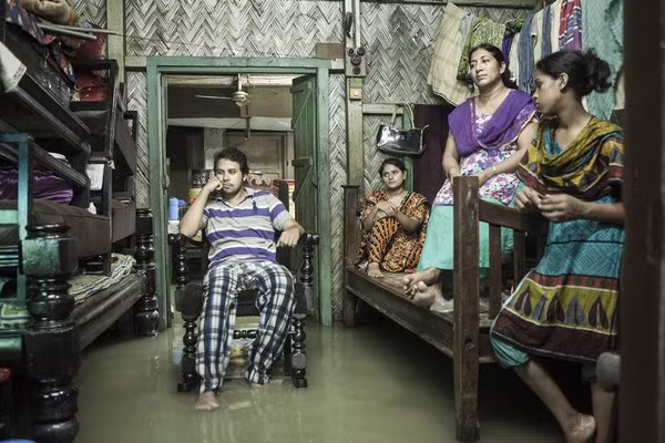 Hình ảnh một gia đình đang xem TV và chờ nước rút ở thành phố Chittagong, Bangladesh. Trong vài năm gần đây, do biến đổi khí hậu nặng nề mà mực nước biển dâng cao - trở thành một vấn đề nan giải tại các khu vực làm ăn và sinh sống của người dân.
