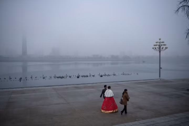 Cặp cô dâu, chú rể đi bộ ven bờ sông Teadong trong một buổi chiều sương giá, phía xa xa là tháp Jechu.
