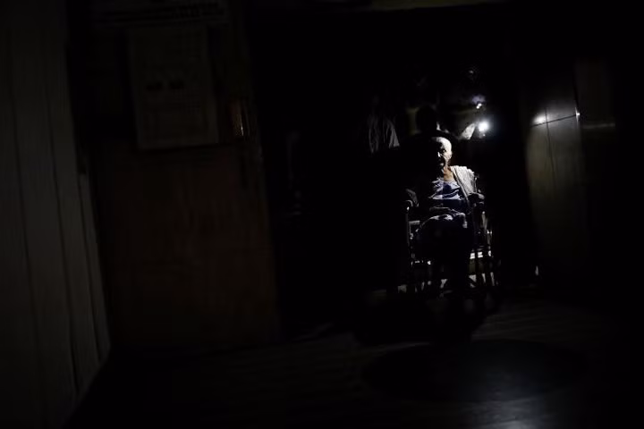 Người dân dùng ánh sáng từ điện thoại di động để giúp bà Olimpia Mora đi xe lăn ra khỏi một tòa nhà lúc bị cúp điện. Venezuela từng giàu có nhờ vào dầu mỏ nhưng giờ lại trong tình trạng thiếu điện nghiêm trọng.