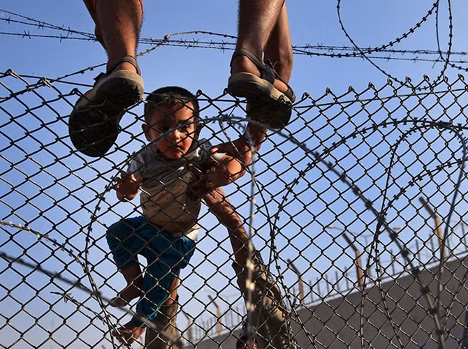 Hàng nghìn người Syria đổ về biên giới với Thổ Nhĩ Kỳ trốn chạy các cuộc giao tranh dữ dội ở miền Bắc và cuộc sống dưới sự kiểm soát của phiến quân IS. Trong ảnh: Một người tị nạn Syria mang con nhỏ vượt rào dây thép gai chạy sang Thổ Nhĩ Kỳ.
